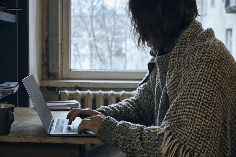 Člověk zabalený v dece pracuje na notebooku u okna s radiátorem